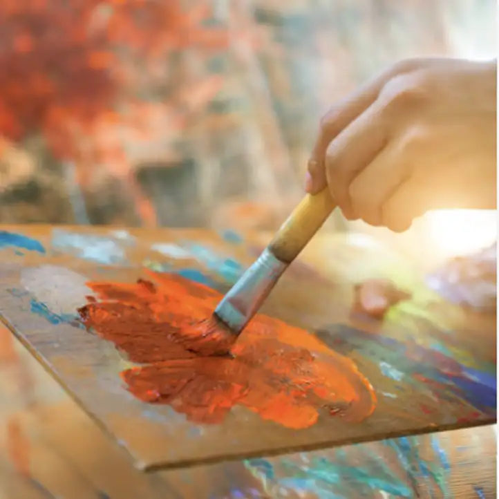 Close-up of artist mixing rich orange paint with brush on palette, showing hands-on process for watercolor and acrylic commissions